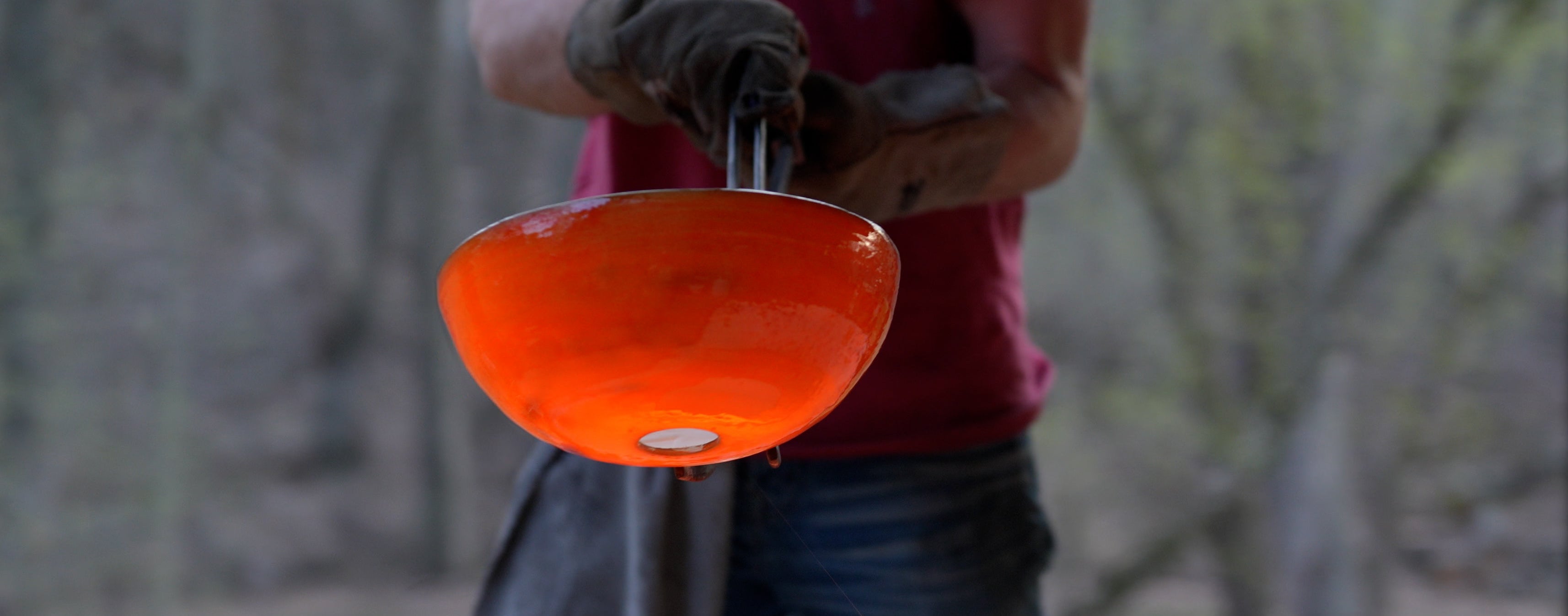 A person in a red shirt and gloves holds a glowing orange molten bowl above a furnace with bright orange liquid, set against an outdoor wooded background.